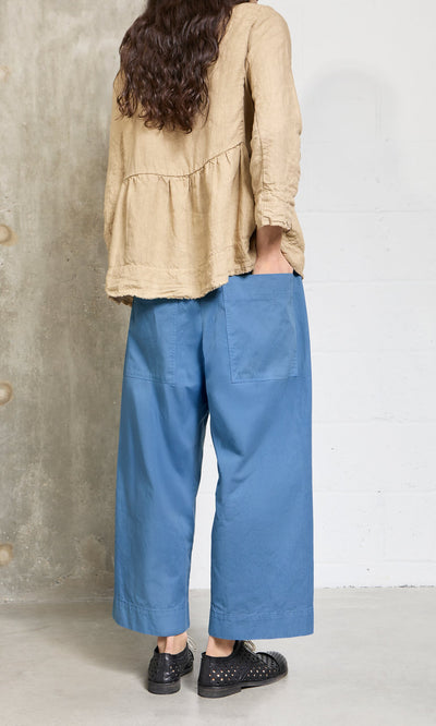 Plümo- Person standing indoors in a beige long-sleeve top and Cromer Pants - French Blue, made in London, shown from the back.
