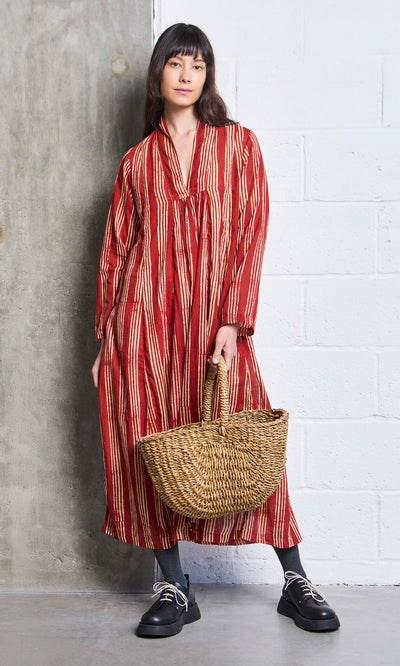 Plümo- Woman in the Lina Stripe Dress holds a woven basket bag, standing by a concrete and white brick wall.