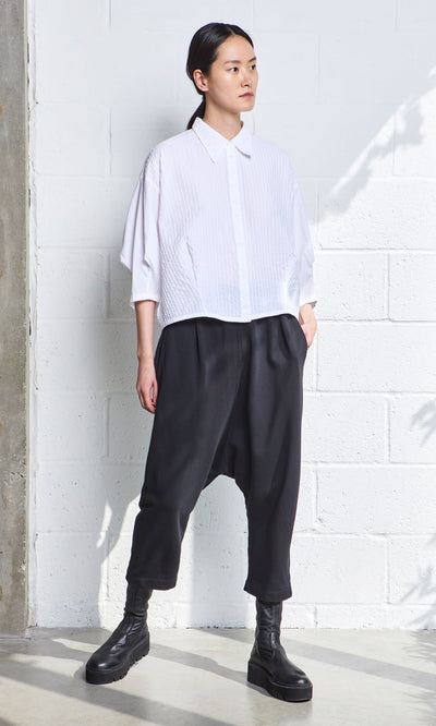 Plümo- Woman stands against a white brick wall, wearing a white shirt and Nerula Pants with black boots.