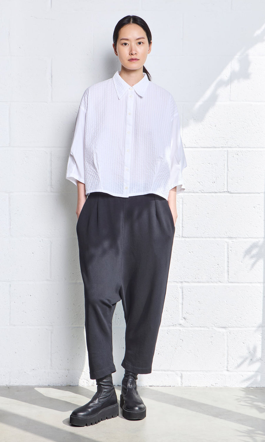 Plümo- Person standing against a white wall in a Fuji Shirt, black drop-crotch pants, and black boots.