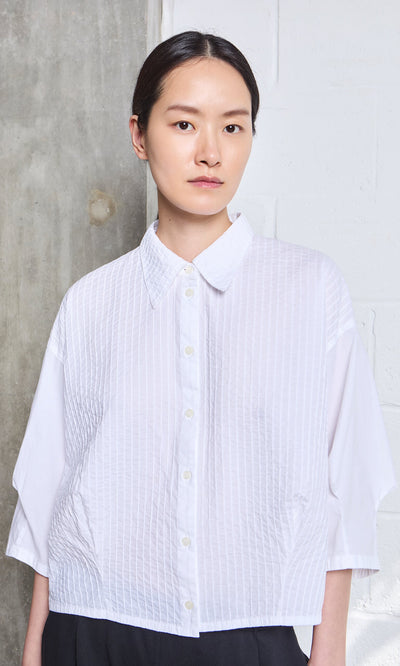 Plümo- Person with straight dark hair wears a white Fuji Shirt, standing against a concrete and white wall.