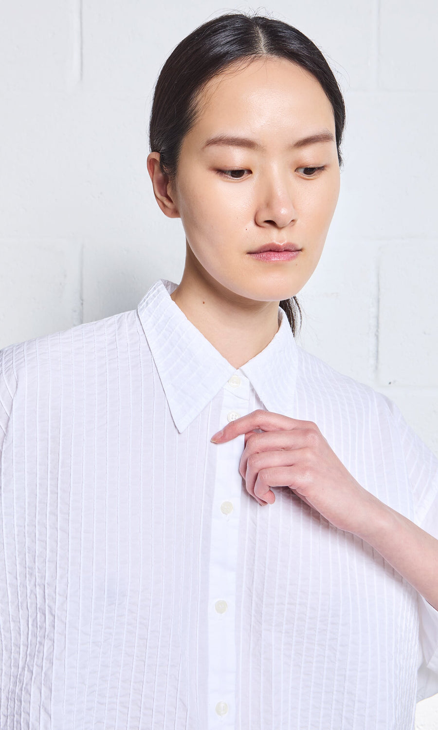 Plümo- Woman wearing the Fuji Shirt by Yacco Maricard, looking down with her hand near her collar against a light background.