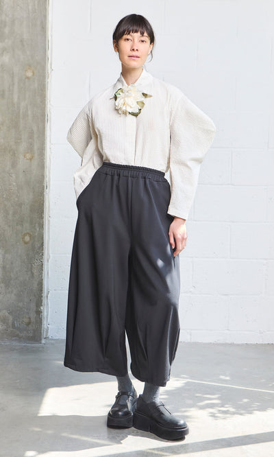 Plümo- Woman in a pintuck cotton Ting Blouse with bishop sleeves, oversized flower brooch, black pants, and black shoes.
