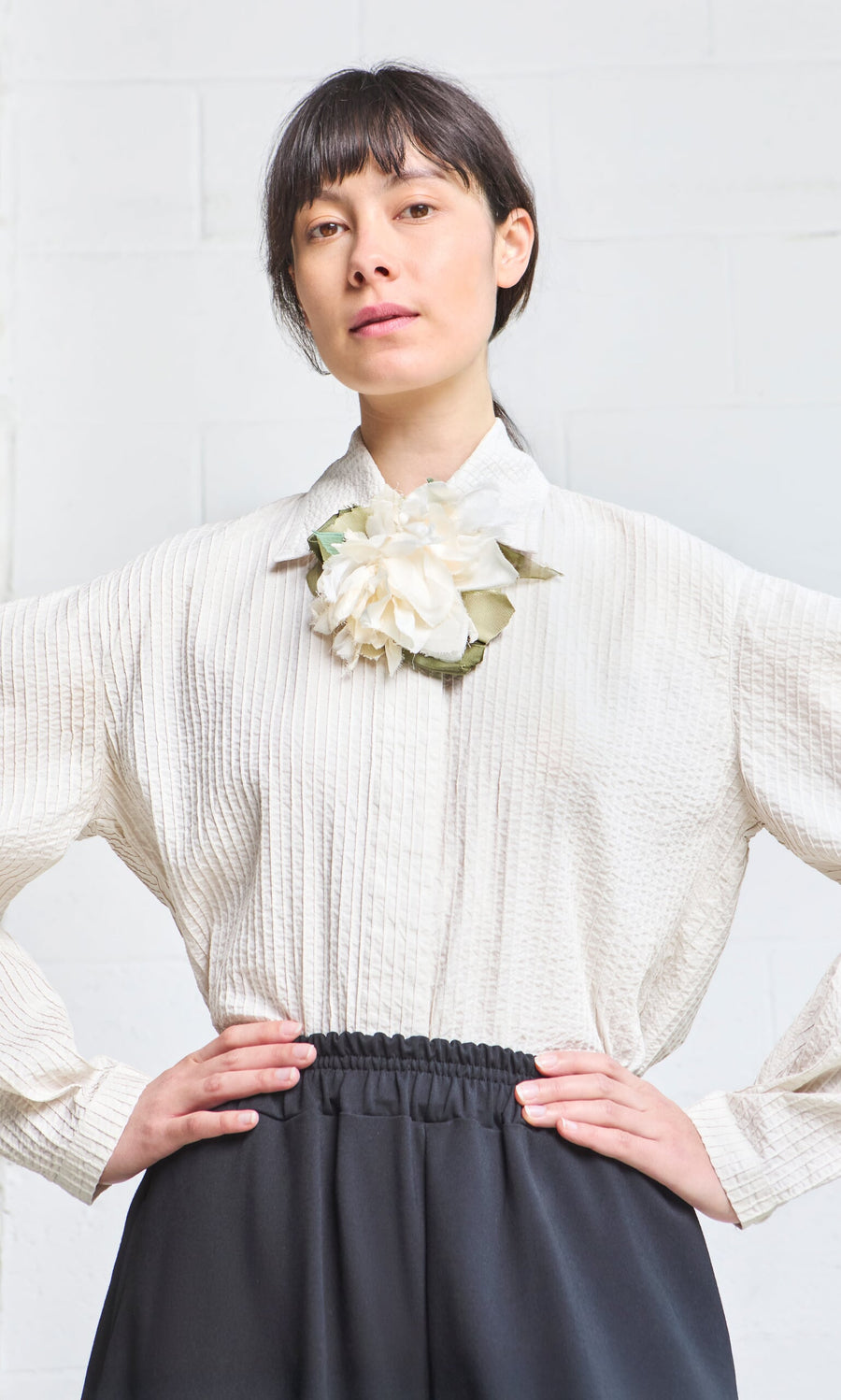Plümo- Woman wearing the Ting Blouse—textured white Japanese shirt with bishop sleeves and a flower brooch—stands hands on hips.