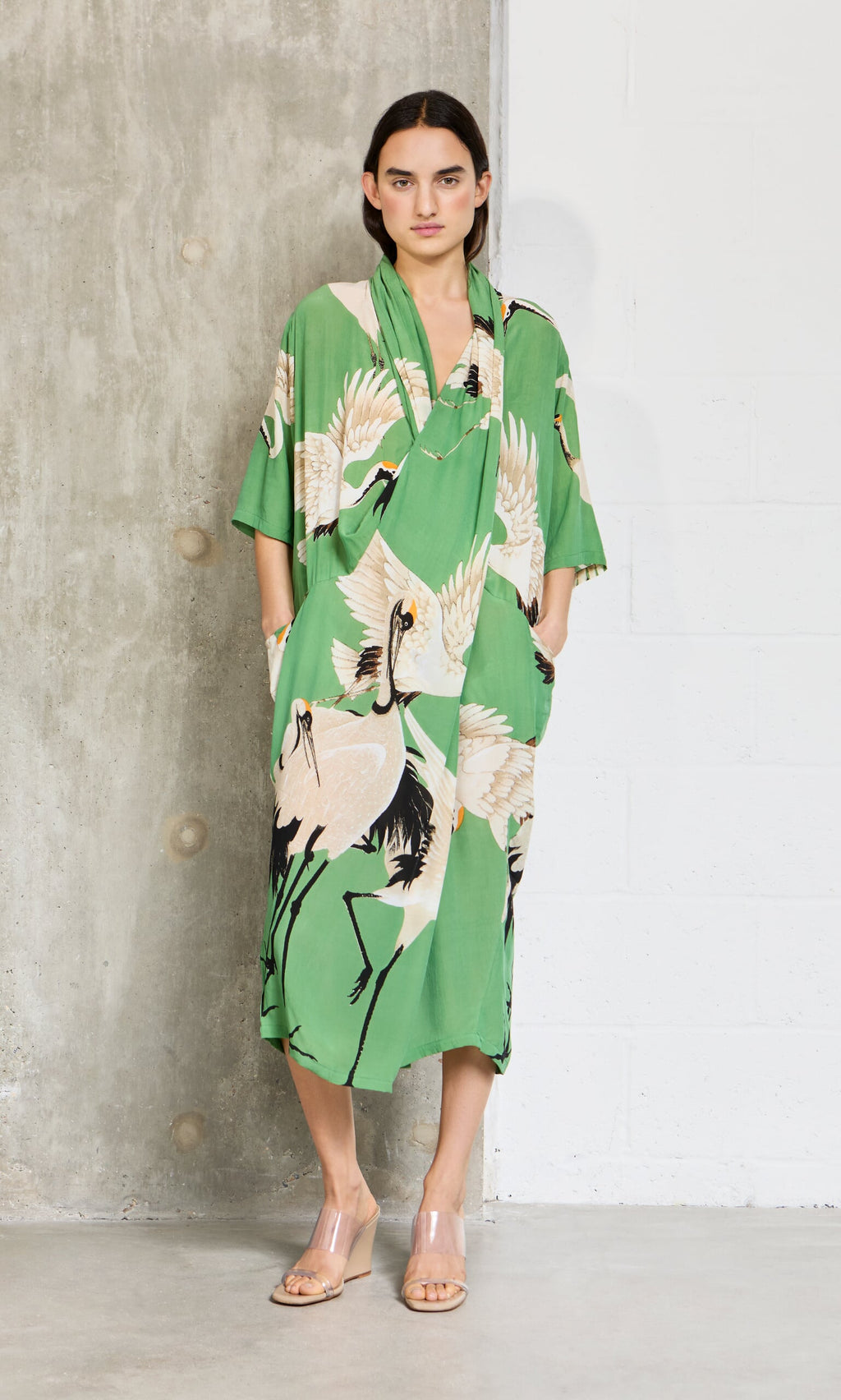 Plümo- Woman wears the Green Stork Dress by a concrete wall, styled with clear heeled sandals.