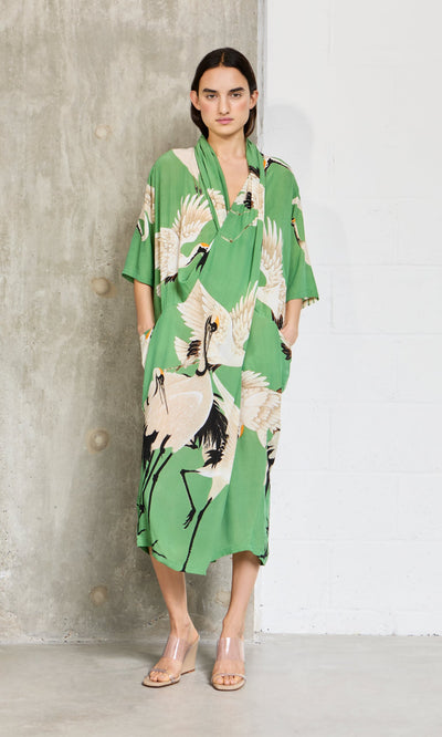 Plümo- Woman wears the Green Stork Dress by a concrete wall, styled with clear heeled sandals.