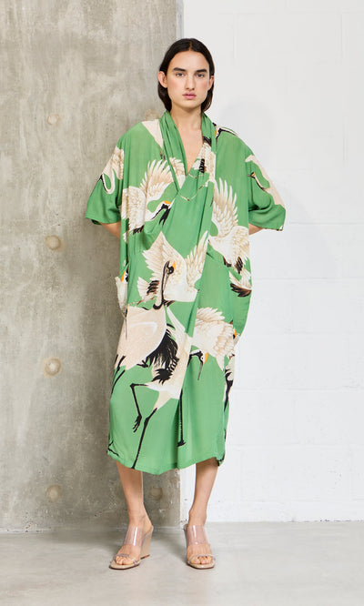 Plümo- Woman wearing the Green Stork Dress stands against a concrete and white wall background.