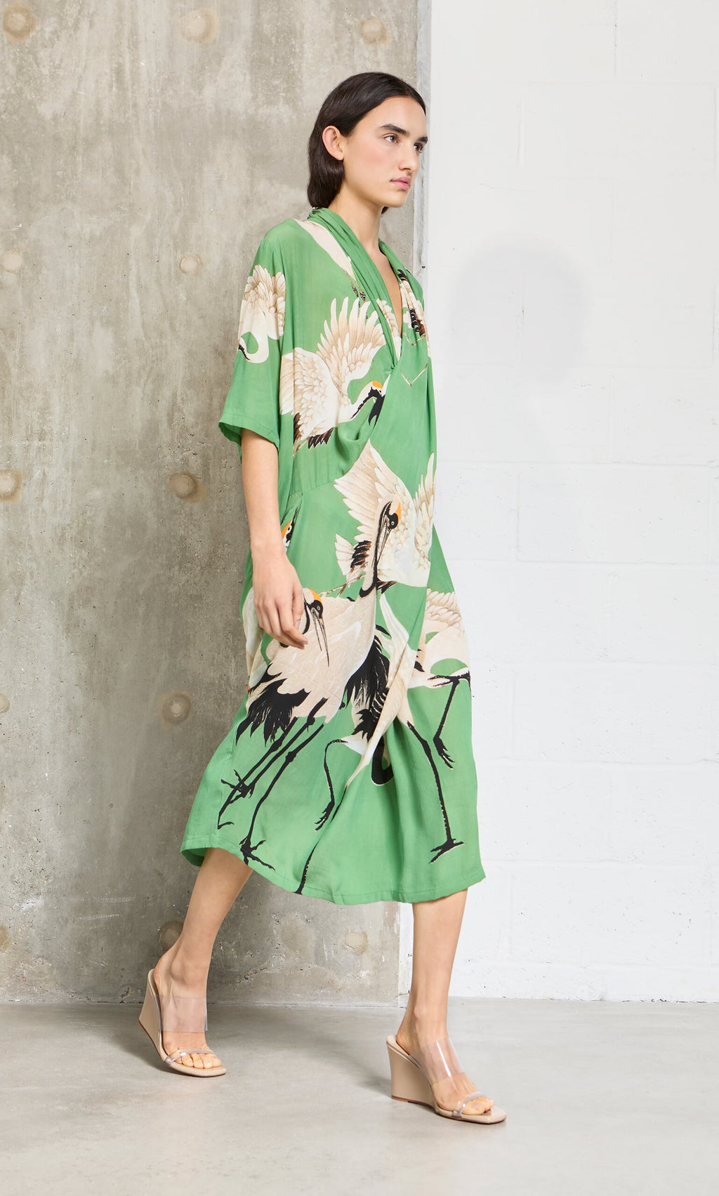 Plümo- Woman in a Green Stork Dress and nude wedge sandals walks by a concrete and white wall.