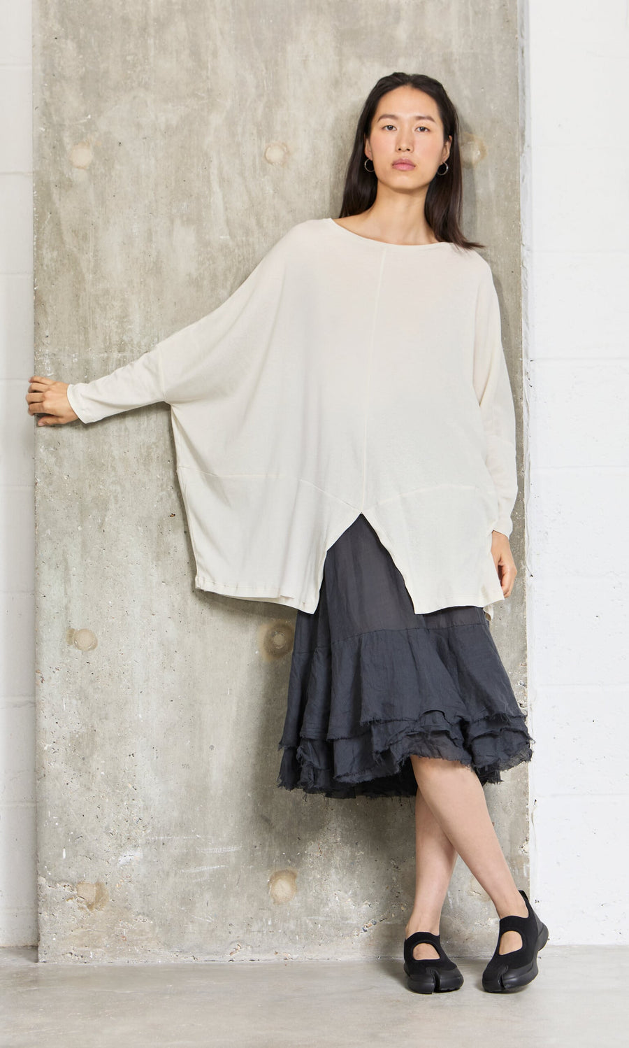 Plümo- Woman stands by a wall wearing the Greige Blouse in soft cotton jersey, a black skirt, and black shoes.