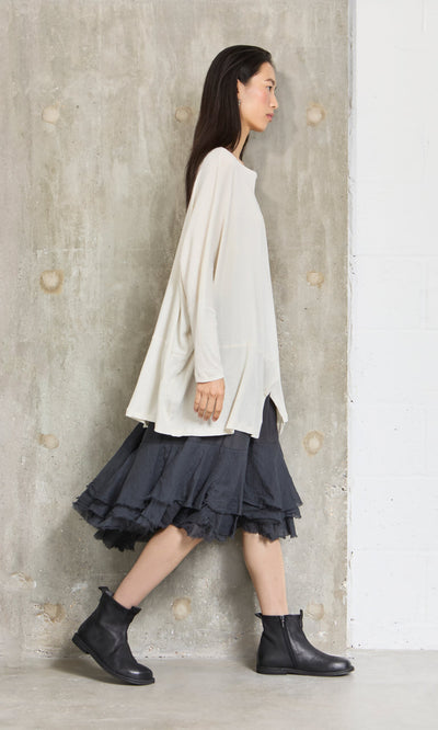 Plümo- Woman in a Greige Blouse, dark layered skirt with zigzag hem, and black ankle boots by a textured concrete wall.