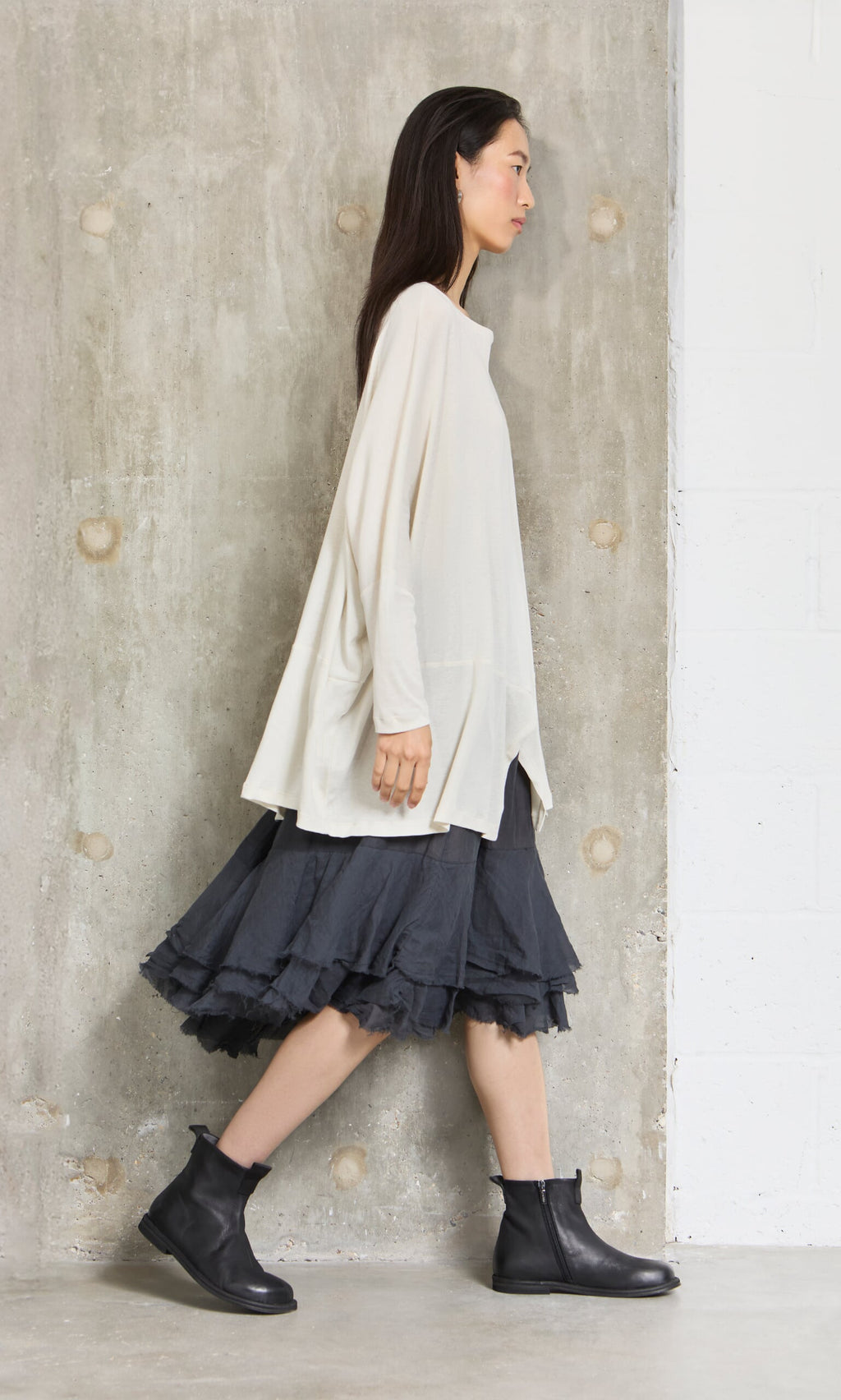 Plümo- Woman in a loose cream top, Nettle Crinkle Skirt, and black boots walks past a textured concrete wall.