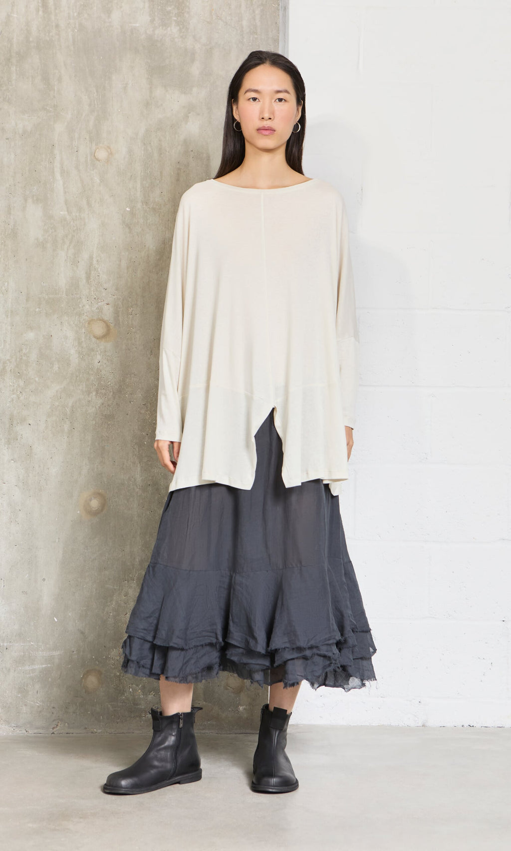 Plümo- Woman stands against a concrete wall, wearing a loose cream top, Nettle Crinkle Skirt, and black ankle boots.