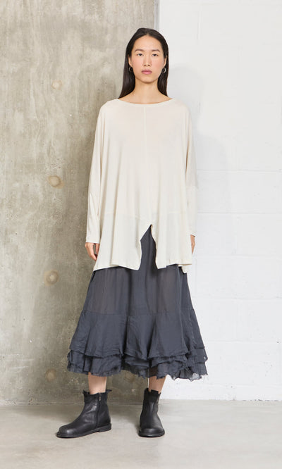 Plümo- Woman stands against a concrete wall, wearing a loose cream top, Nettle Crinkle Skirt, and black ankle boots.