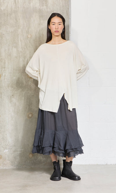 Plümo- Woman indoors in a Greige Blouse, black ruffled skirt with zigzag hem, and black ankle boots.