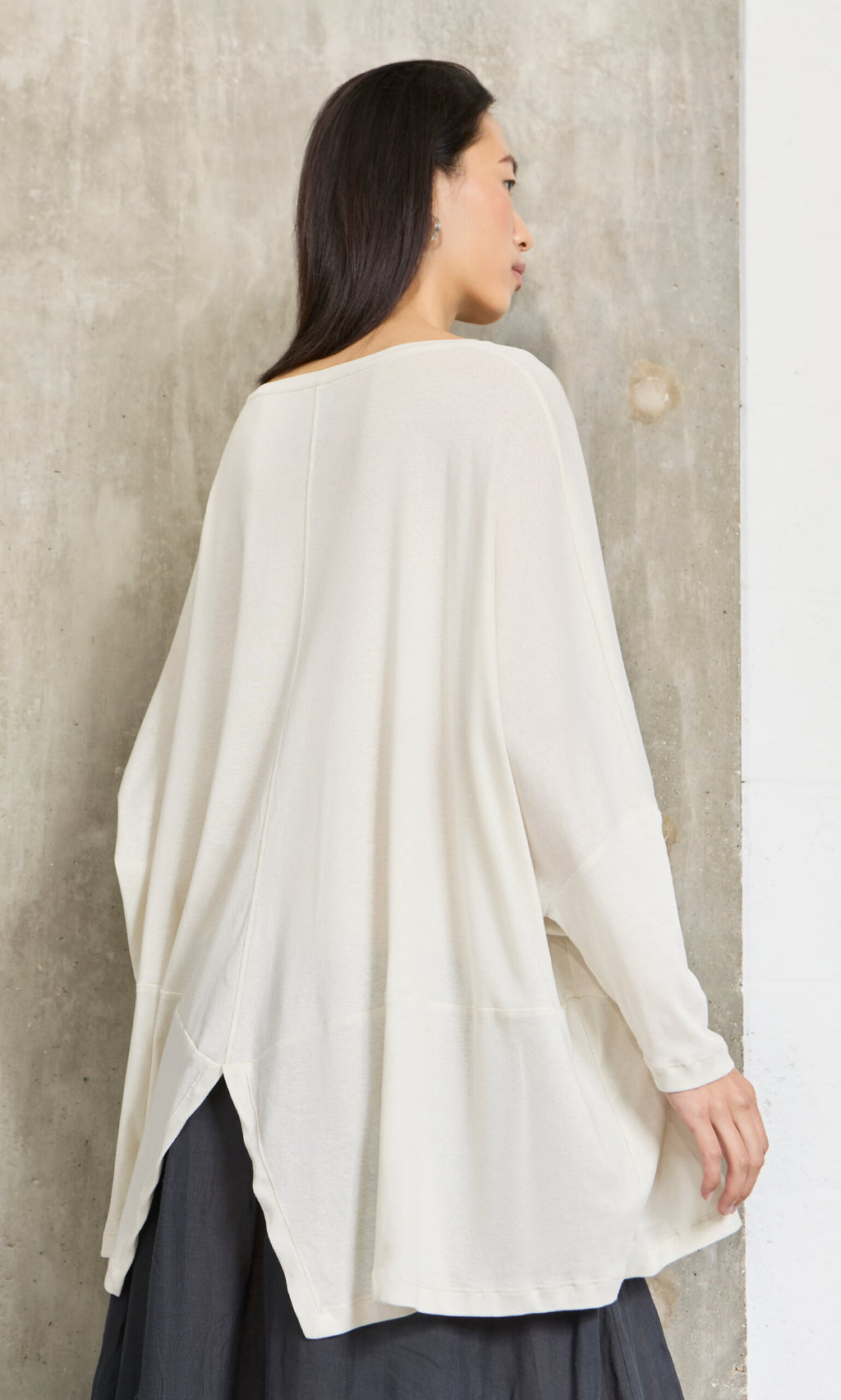 Plümo- Woman in a loose, long-sleeved Greige Blouse with split back, posed against a textured concrete wall.