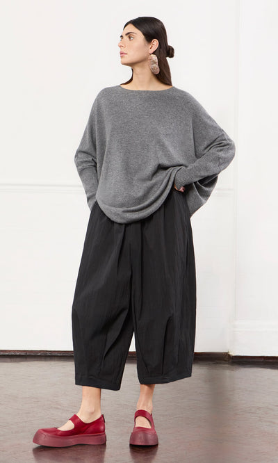 Plümo- A woman in a Grey Cashmere Jumper with an oversized fit and red shoes stands indoors, looking to the side.
