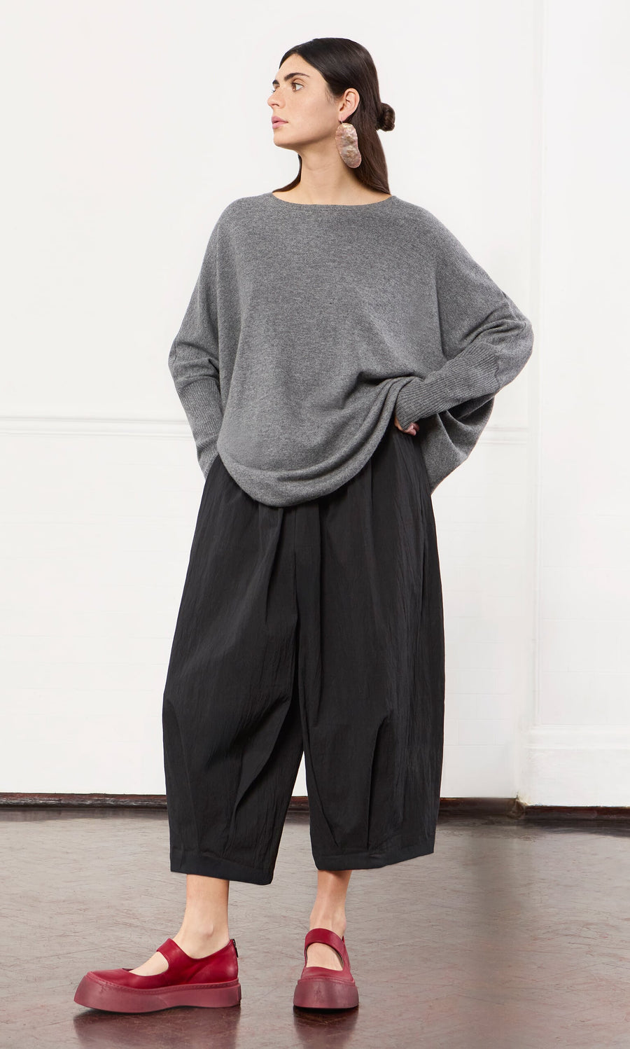 Plümo- A woman in a Grey Cashmere Jumper with an oversized fit and red shoes stands indoors, looking to the side.