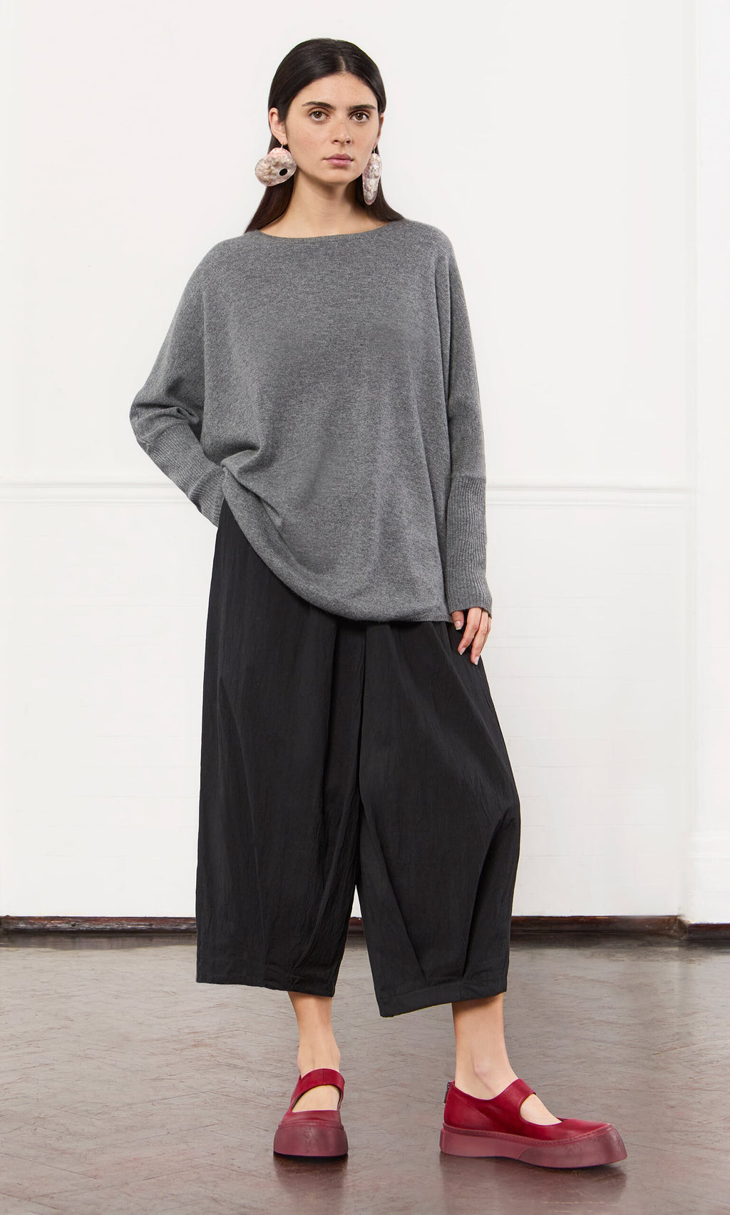 Plümo- Woman in a Grey Cashmere Jumper, wide black pants, and red shoes stands against a white wall.