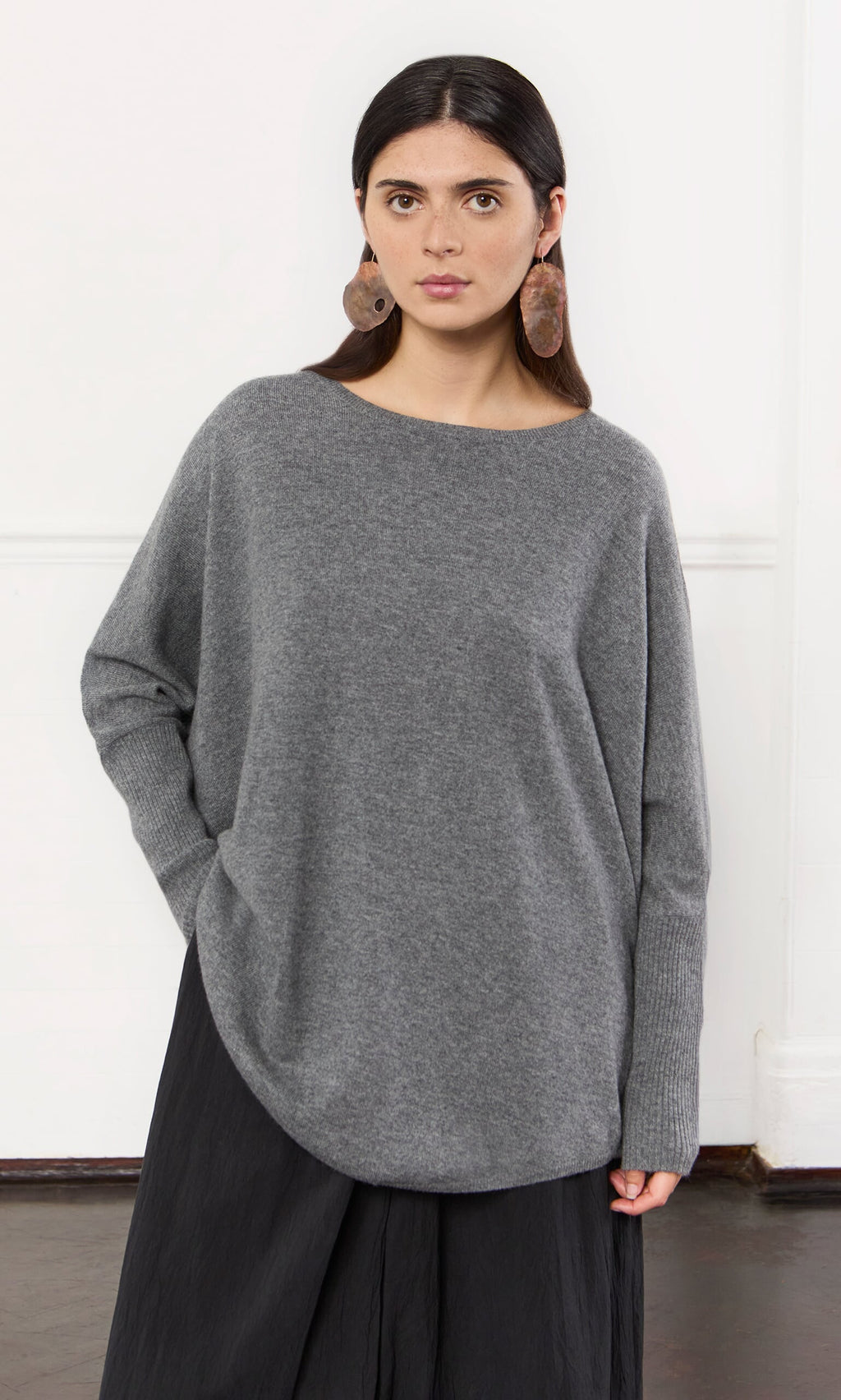 Plümo- Woman with long dark hair in a grey cashmere jumper, black skirt, and brown earrings, standing indoors.