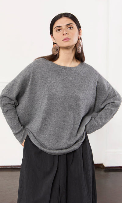 Plümo- Woman wears a Grey Cashmere Jumper, black skirt, and large round earrings, standing with hands in pockets.