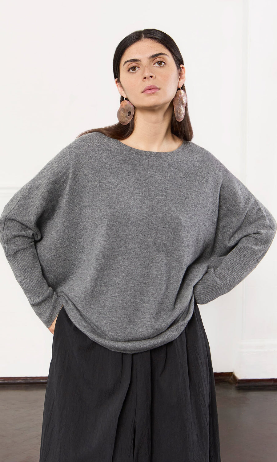 Plümo- Woman wears a Grey Cashmere Jumper, black skirt, and large round earrings, standing with hands in pockets.