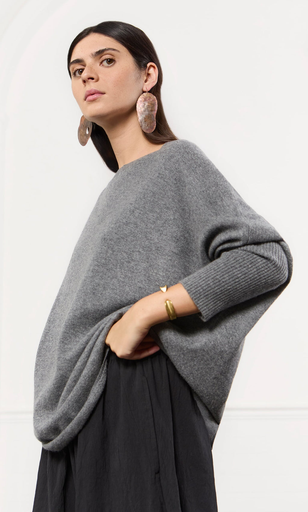 Plümo- Woman in an oversized grey cashmere jumper and black skirt, posing confidently with bold earrings and a gold bracelet.