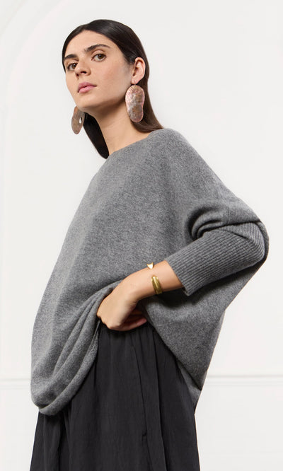Plümo- Woman in an oversized grey cashmere jumper and black skirt, posing confidently with bold earrings and a gold bracelet.