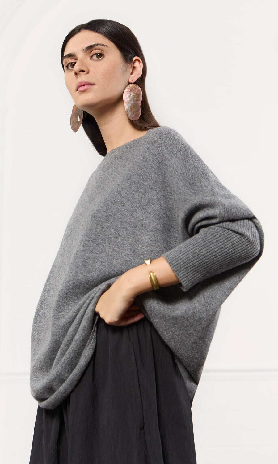 Plümo- Woman in an oversized grey cashmere jumper and black skirt, posing confidently with bold earrings and a gold bracelet.