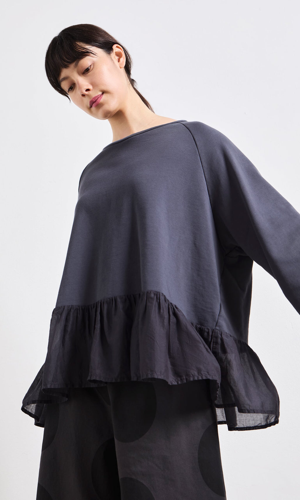 Plümo- Woman wearing the Grey Ruffle Top with wide sleeves and peplum ruffles, looking downward against a white background.