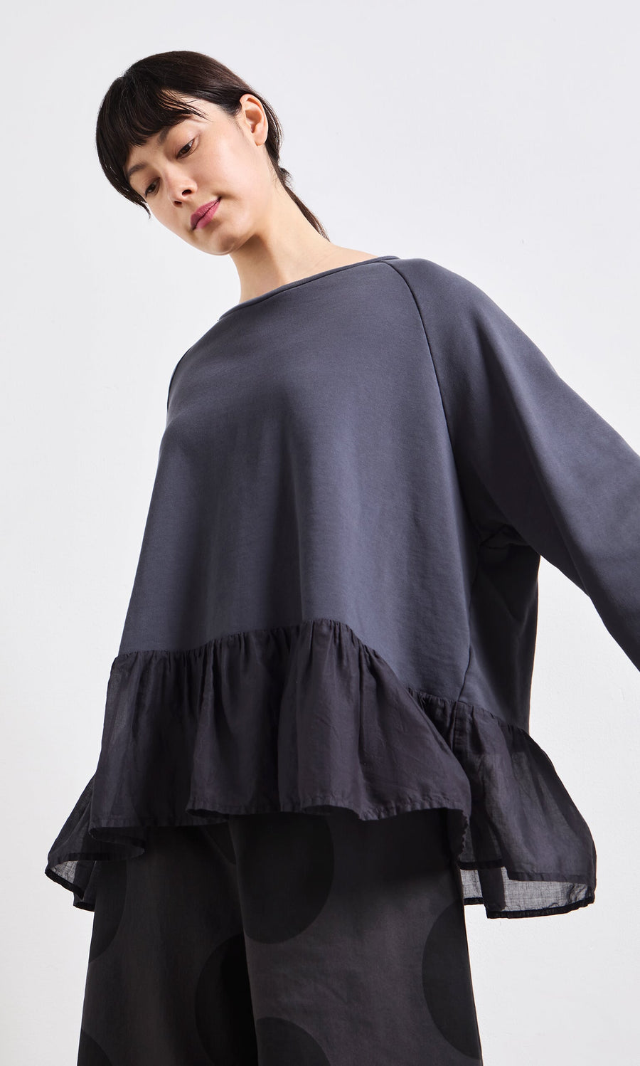 Plümo- Woman wearing the Grey Ruffle Top with wide sleeves and peplum ruffles, looking downward against a white background.