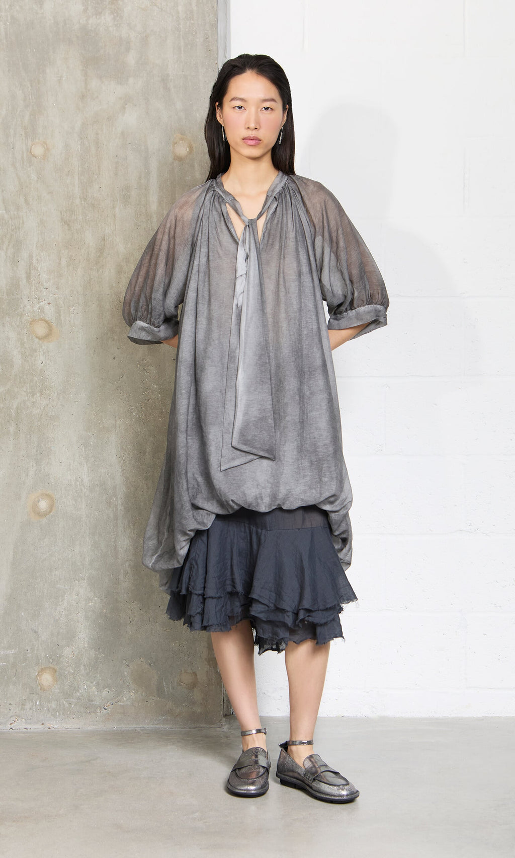 Plümo- Woman in a Grey Voile Blouse with puffy sleeves over a ruffled black skirt stands against a concrete wall.