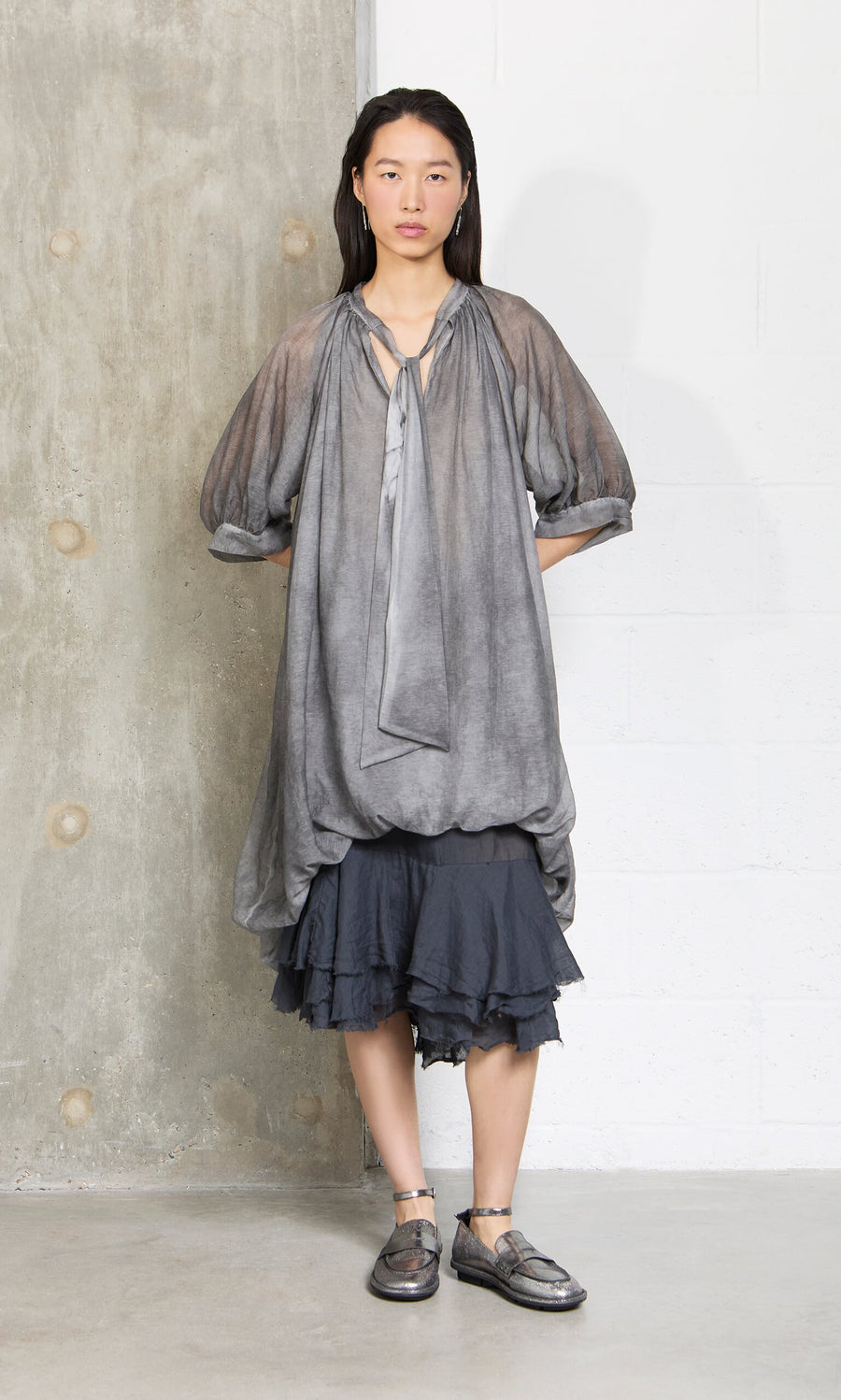 Plümo- Woman in a Grey Voile Blouse with puffy sleeves over a ruffled black skirt stands against a concrete wall.