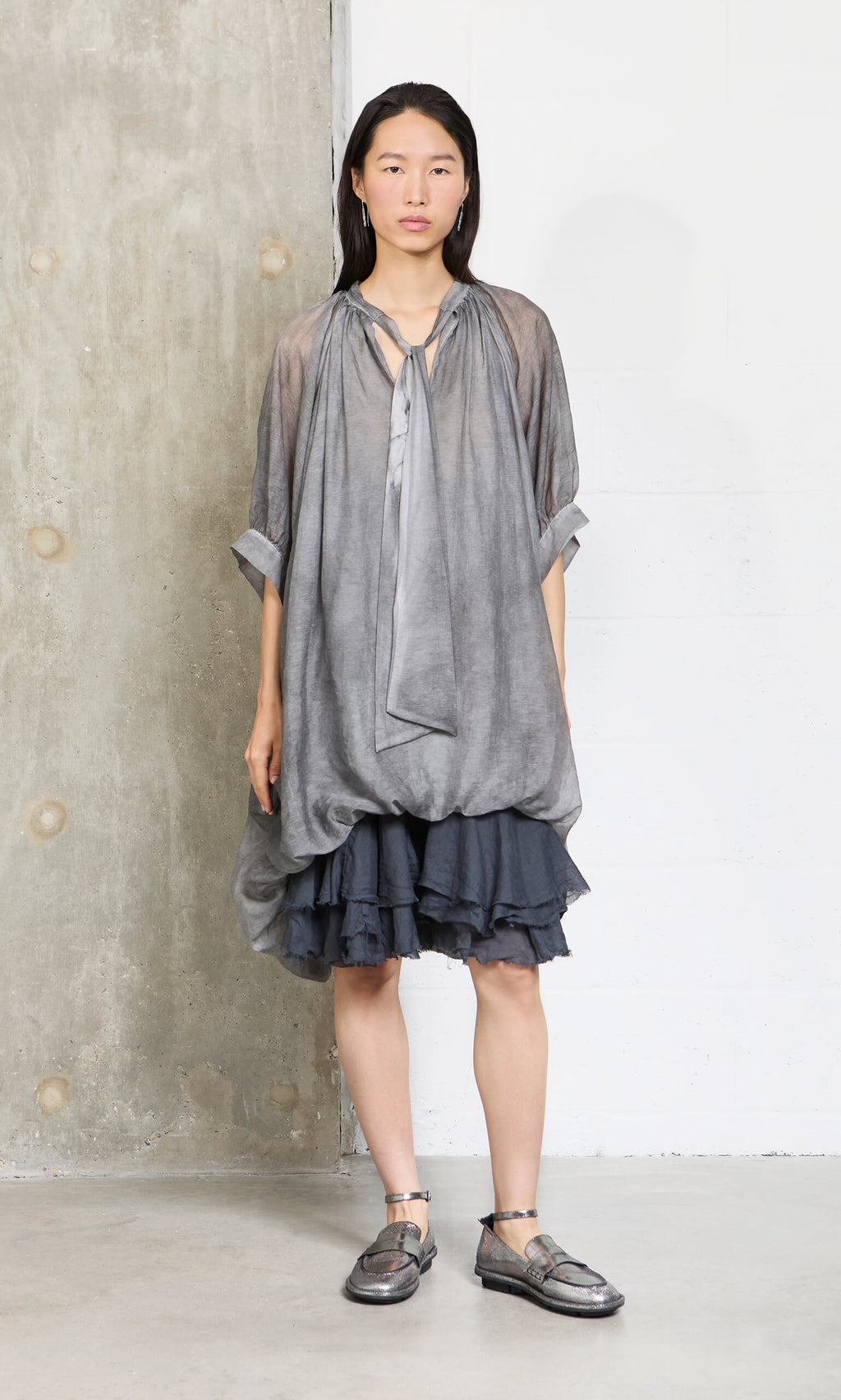 Plümo- A woman in a Grey Voile Blouse and metallic shoes stands by a concrete and white wall, looking ahead.