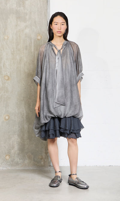 Plümo- A woman in a Grey Voile Blouse and metallic shoes stands by a concrete and white wall, looking ahead.