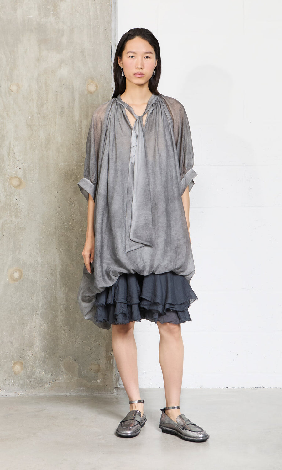 Plümo- A woman in a Grey Voile Blouse and metallic shoes stands by a concrete and white wall, looking ahead.