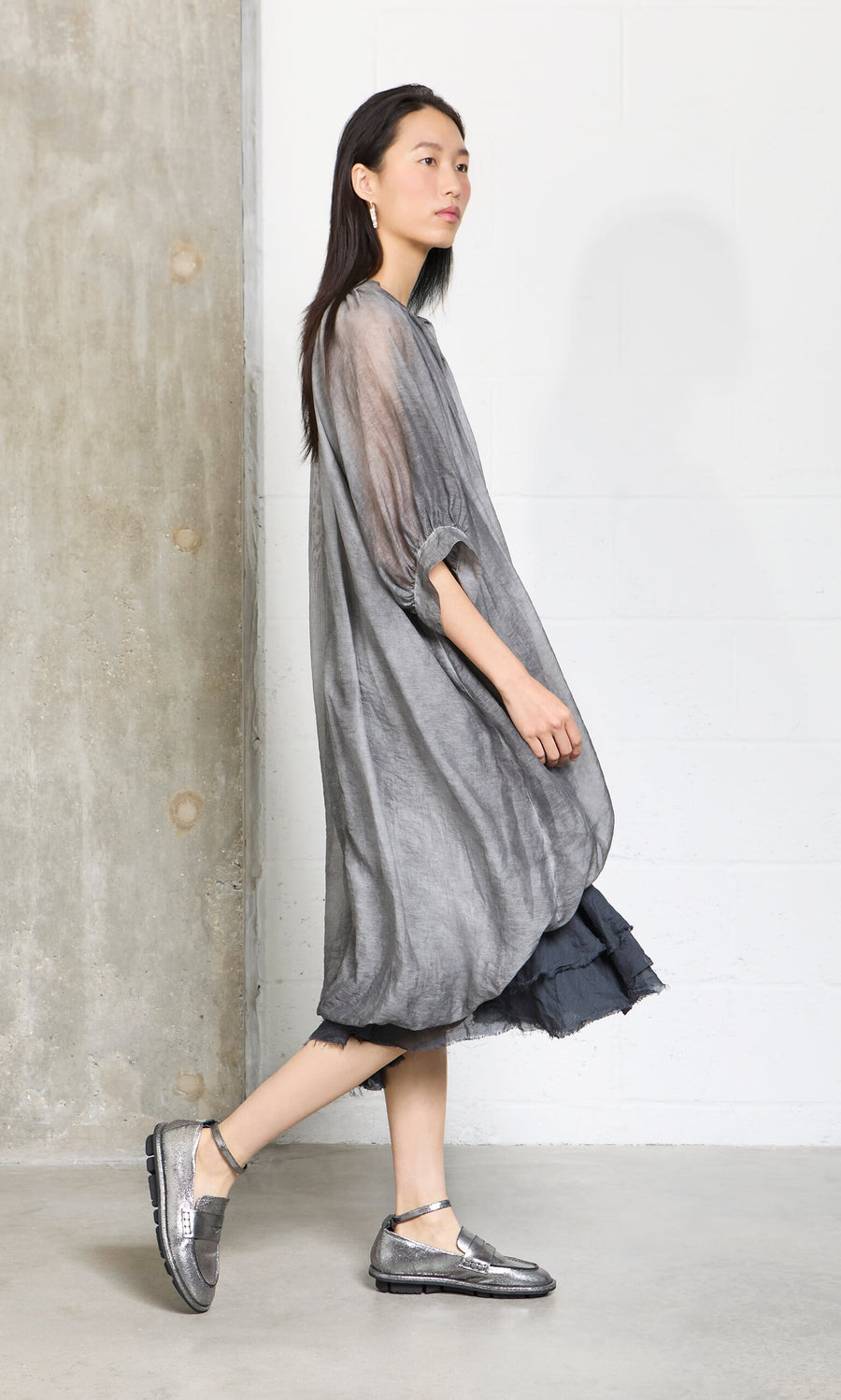 Plümo- A woman in a Grey Voile Blouse and metallic shoes walks indoors against concrete and white walls.