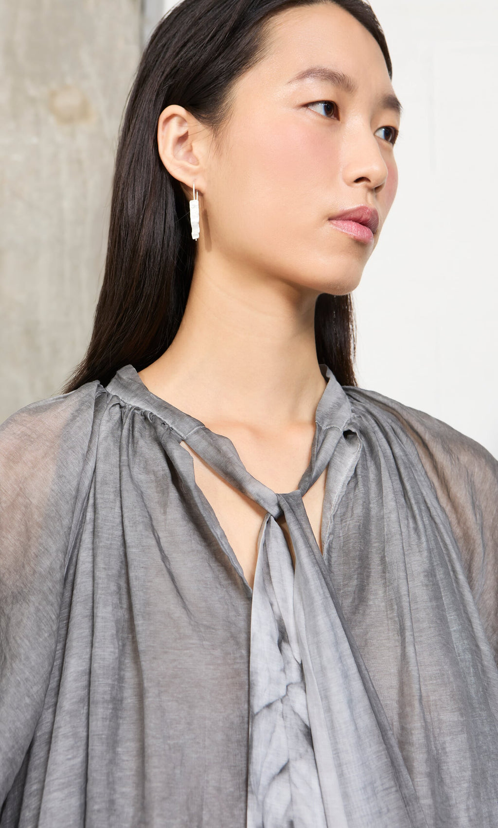 Plümo- Woman with long dark hair wears a Grey Voile Blouse and rectangular drop earrings, looking off to the side.
