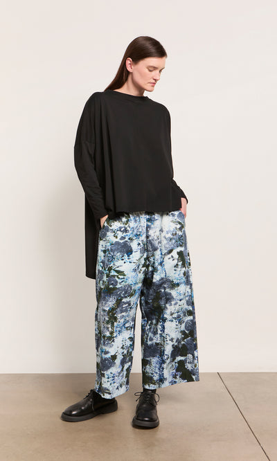 Plümo  Person in an oversized Hansen Tee and wide-leg, blue-patterned pants stands against a light wall, hands in pockets.