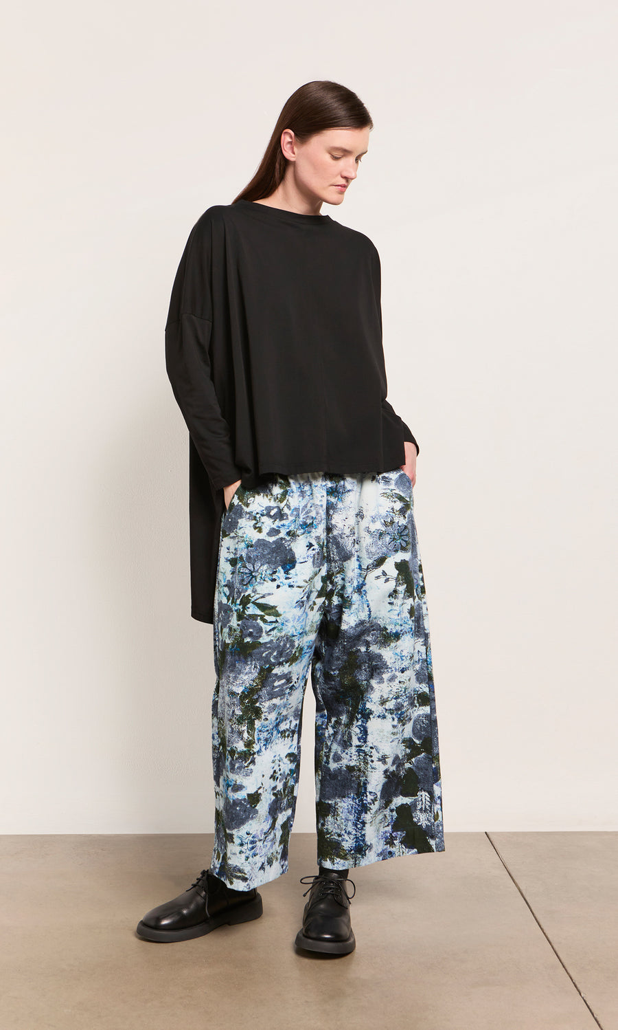 Plümo  Person in an oversized Hansen Tee and wide-leg, blue-patterned pants stands against a light wall, hands in pockets.