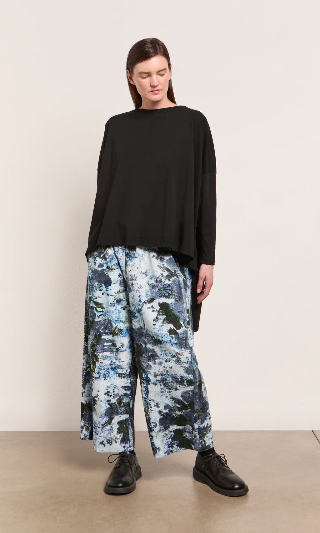 Plümo  Woman in Caccia Pants—wide-leg cotton with wintery print—and a loose black top stands by a plain wall.