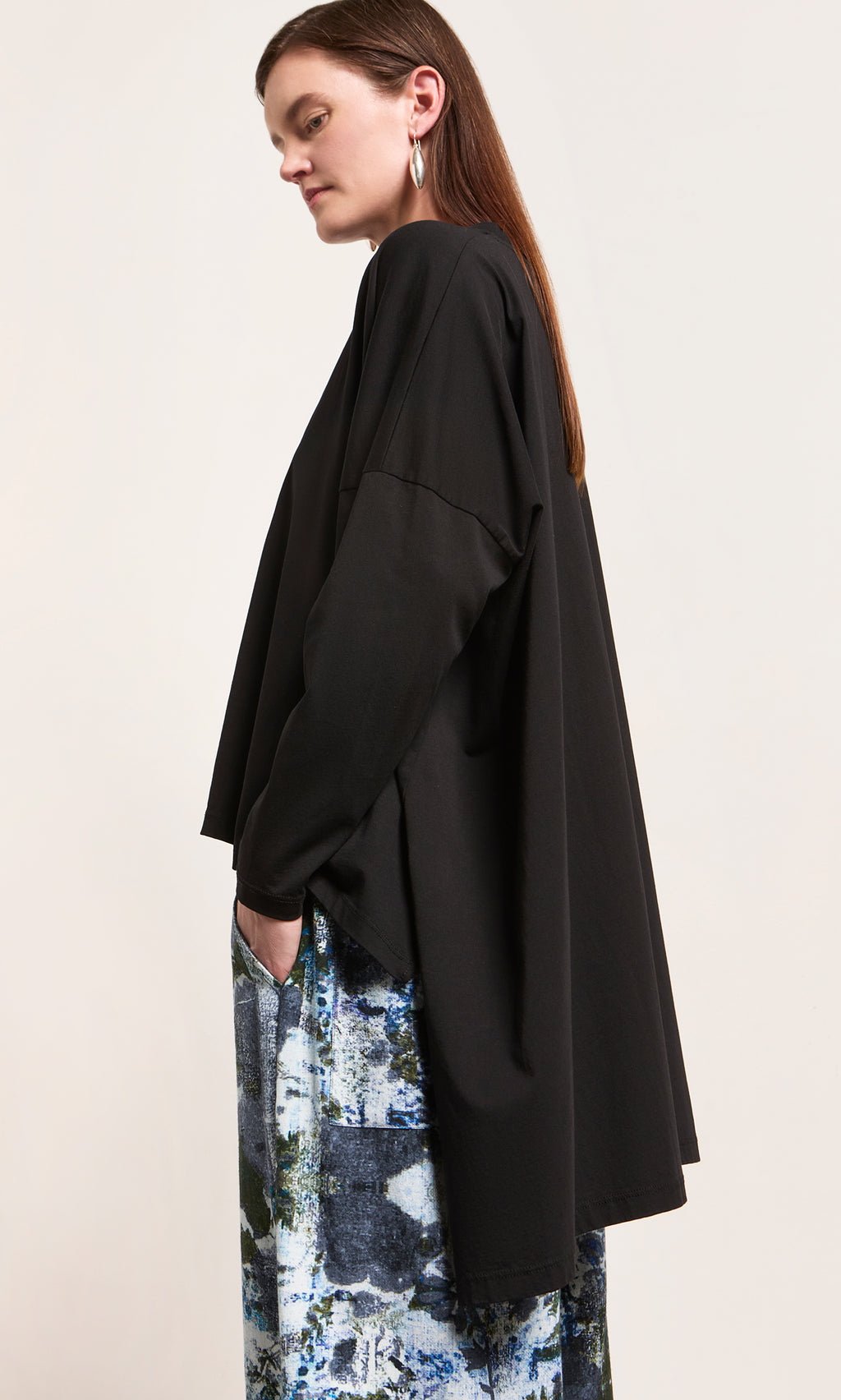 Plümo  Woman in an oversized Hansen Tee and patterned blue pants, standing sideways with her hand in a pocket.