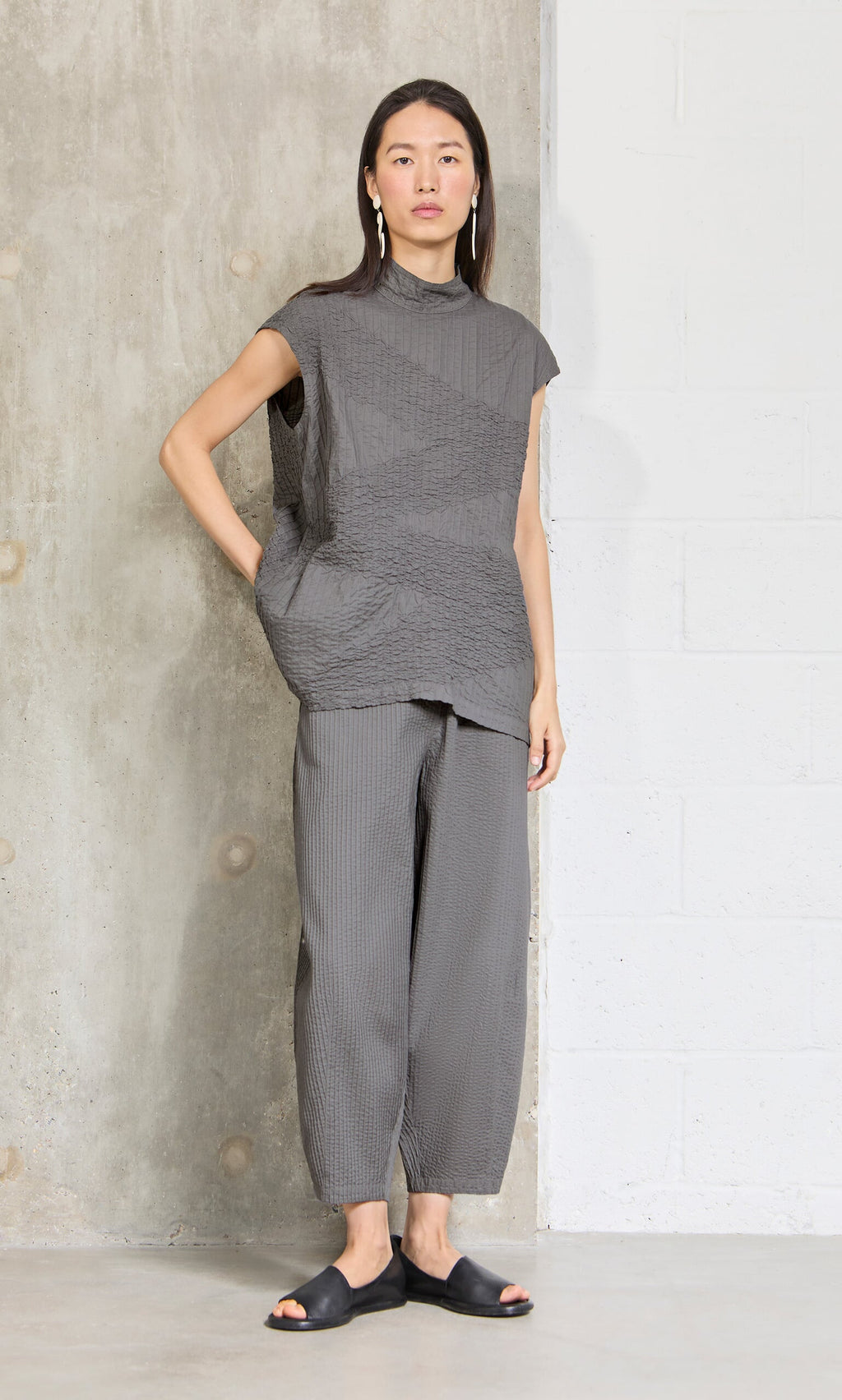 Plümo- Woman stands against a wall in a loose gray top, Haru Grey Pants, black slides, and dangling earrings.