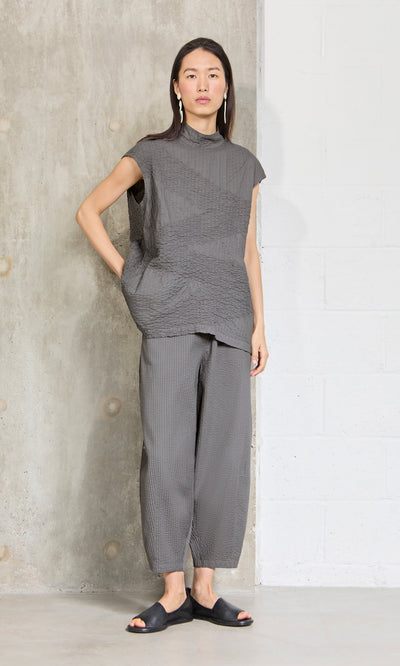 Plümo- Woman stands against a wall in a loose gray top, Haru Grey Pants, black slides, and dangling earrings.