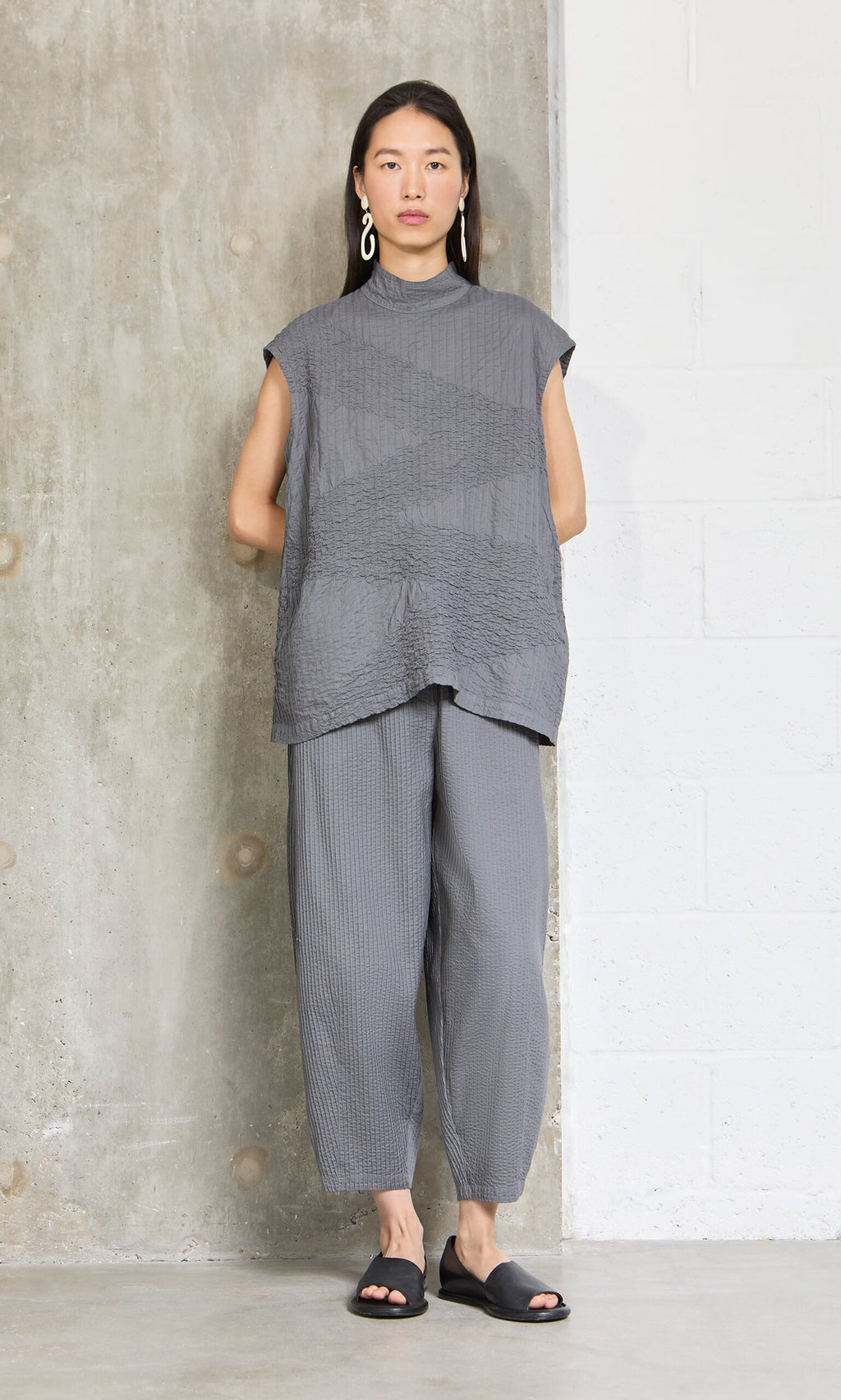 Plümo- Woman stands against a concrete wall wearing the Minato Top, matching pants, black sandals, and large earrings.