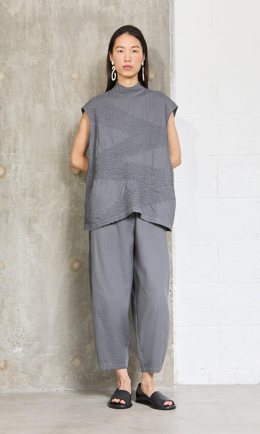 Plümo- Woman stands against a concrete wall wearing the Minato Top, matching pants, black sandals, and large earrings.