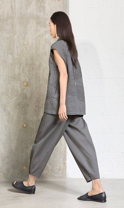 Plümo- Woman in Haru Grey Pants and a loose gray top, black flats, walking indoors by concrete and white walls, side view.
