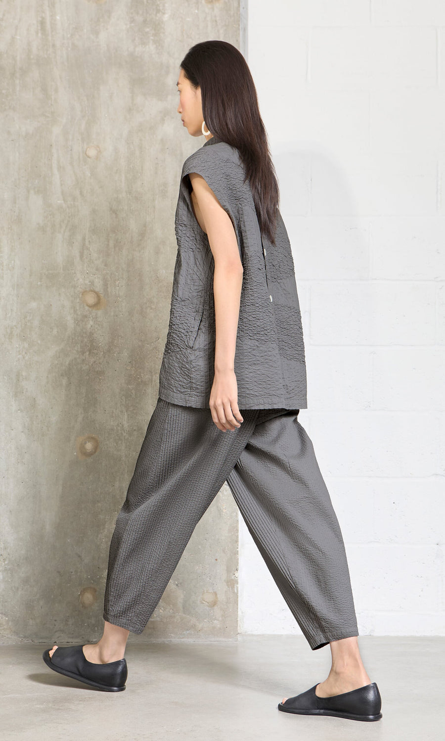 Plümo- Woman in Haru Grey Pants and a loose gray top, black flats, walking indoors by concrete and white walls, side view.
