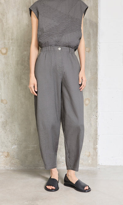 Plümo- Person standing by a concrete wall in Haru Grey Pants, gray top, and black open-toe sandals.