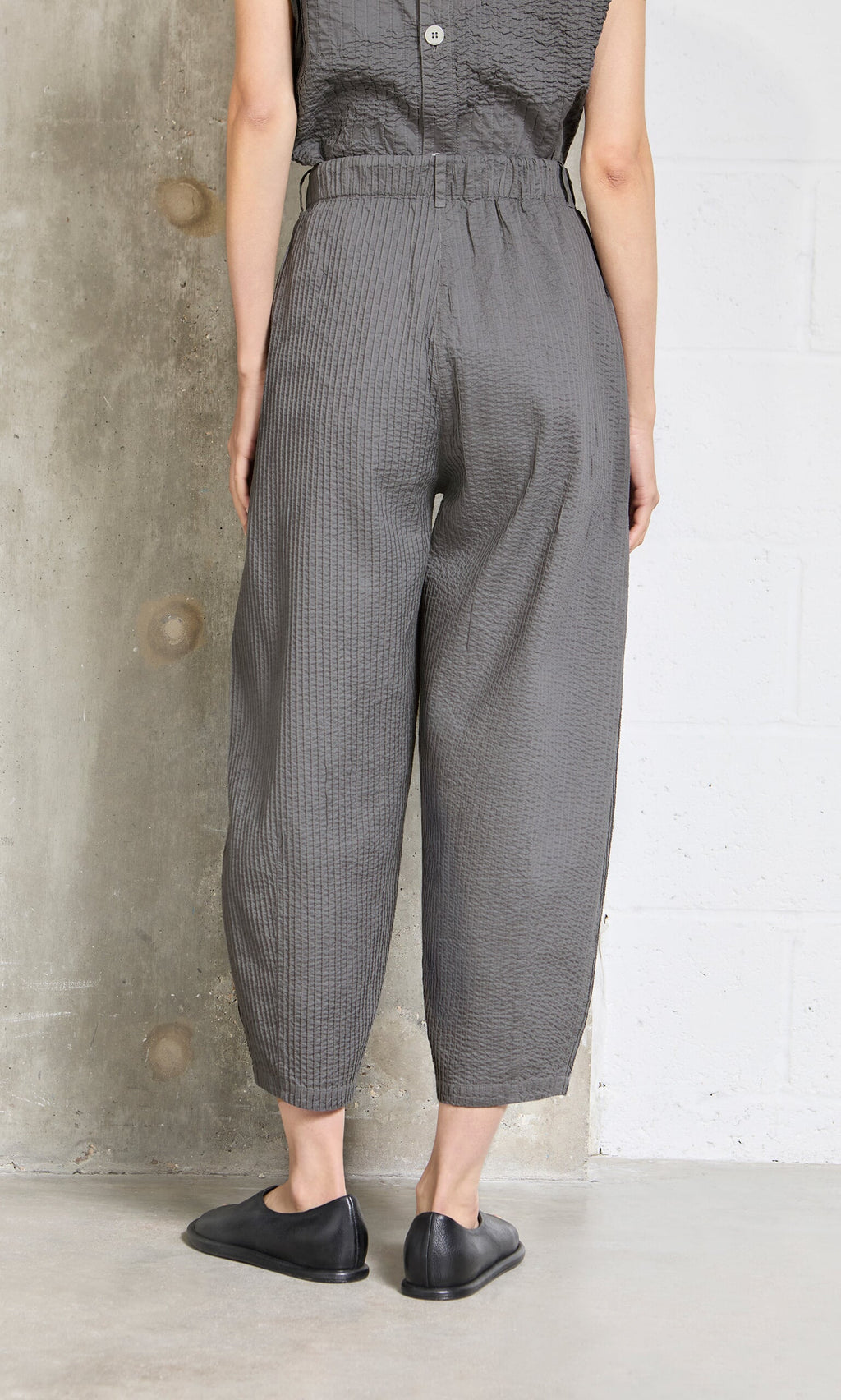 Plümo- Person wearing loose Haru Grey Pants and black shoes, standing with their back to a concrete wall.