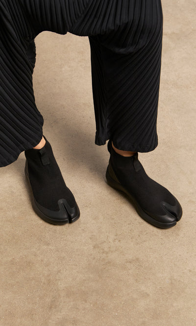 Plümo  Person wearing Higher Tabi Boots and pleated black pants, standing on a tan floor.